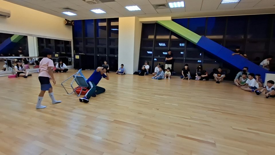 教練在活動室示範用折疊椅擋在身前，孩子們沿牆坐著看