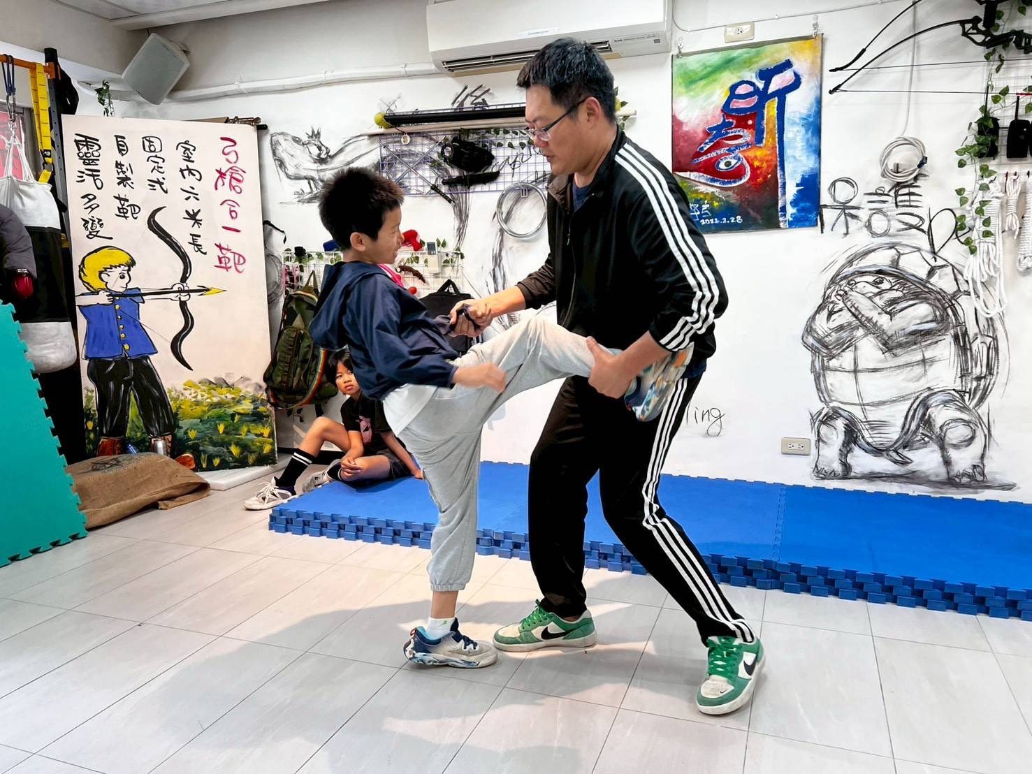 教練一手接住男孩踢出的腳、一手牽著他的手，男孩笑著練習踢擊