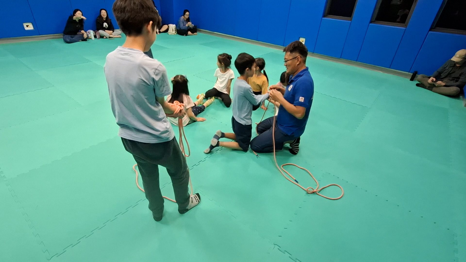 教練跪在地墊上和孩子一起拉繩子打結，家長坐在旁邊看