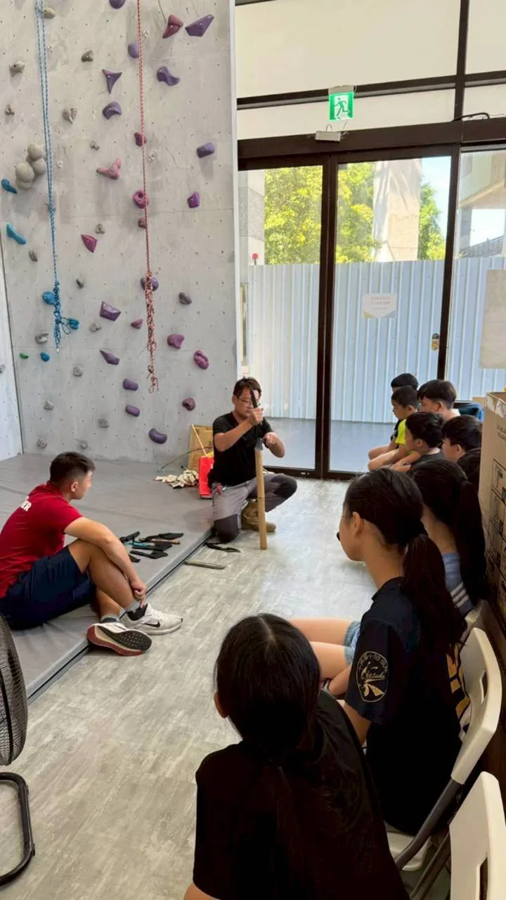 攀岩館裡，教練蹲著示範野外求生工具的用法，學生在旁邊看